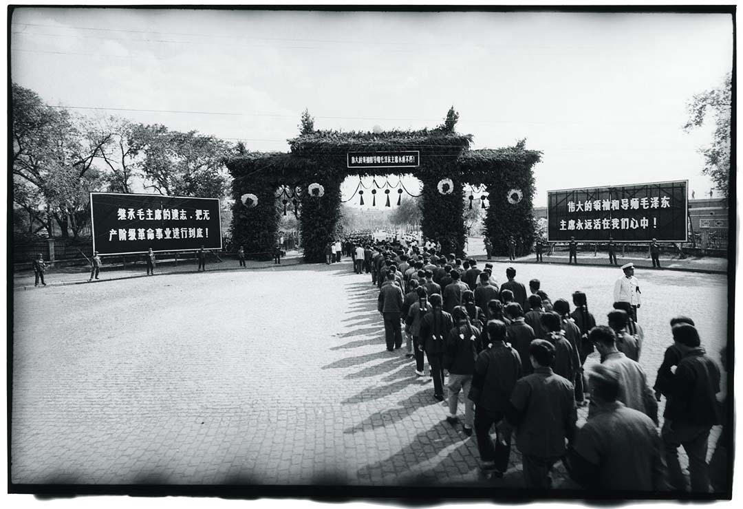 1976年9月18日,哈爾濱,人民前往廣場參加當地悼念毛澤東的儀式。廣場大門前的告示版上寫有:「繼承毛主席的遺志,把無產階級革命事業進行到底!」以及「偉大的領袖和導師毛澤東主席永遠活在我們心中!」。