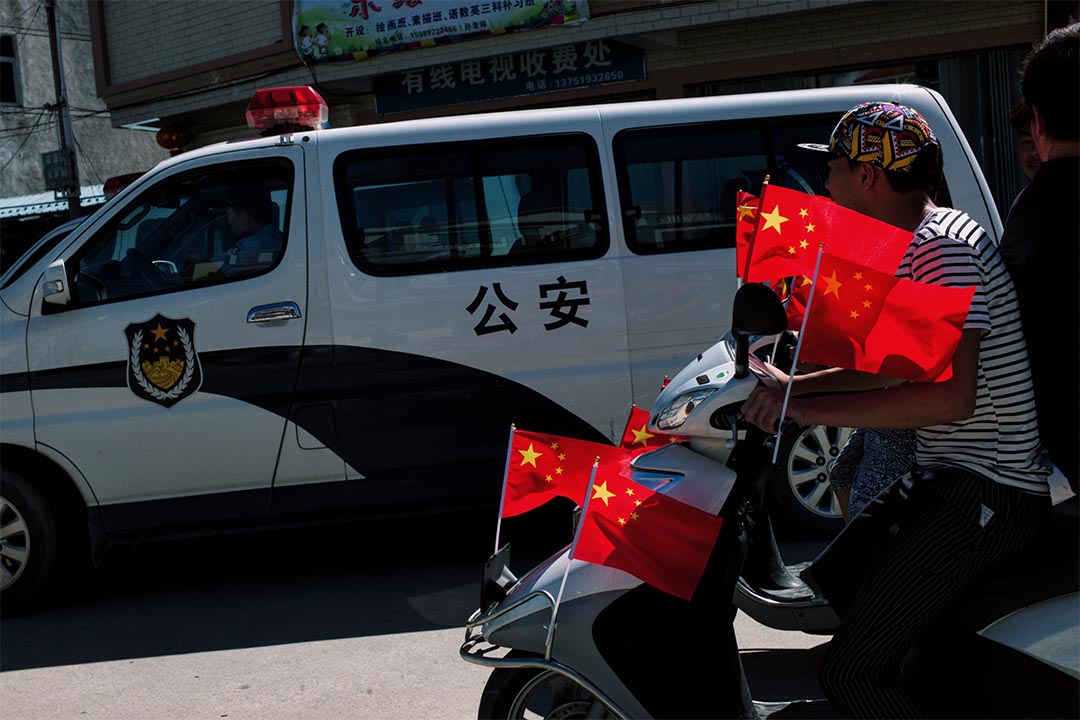 參與遊行的村民乘著插滿國旗的摩托車,駛經停泊於村口的公安車輛。