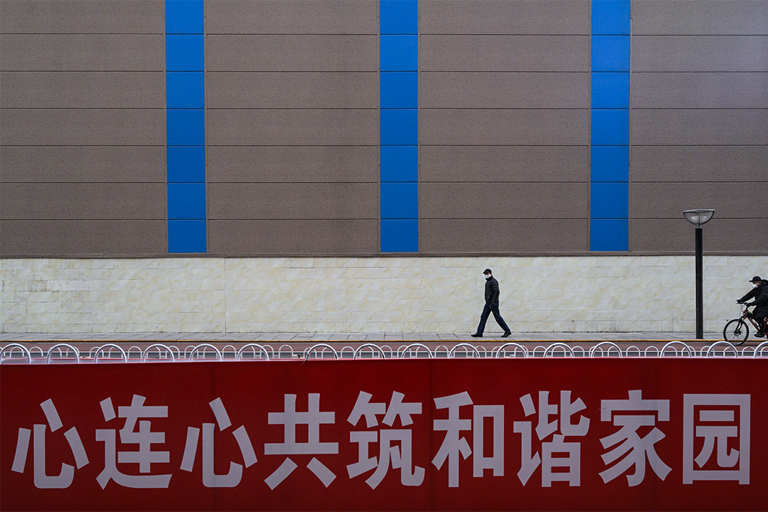 2020年2月18日中國北京,戴著口罩的男子在商業街上走過,一宣傳橫幅上寫著“心連心共築和諧家園”。