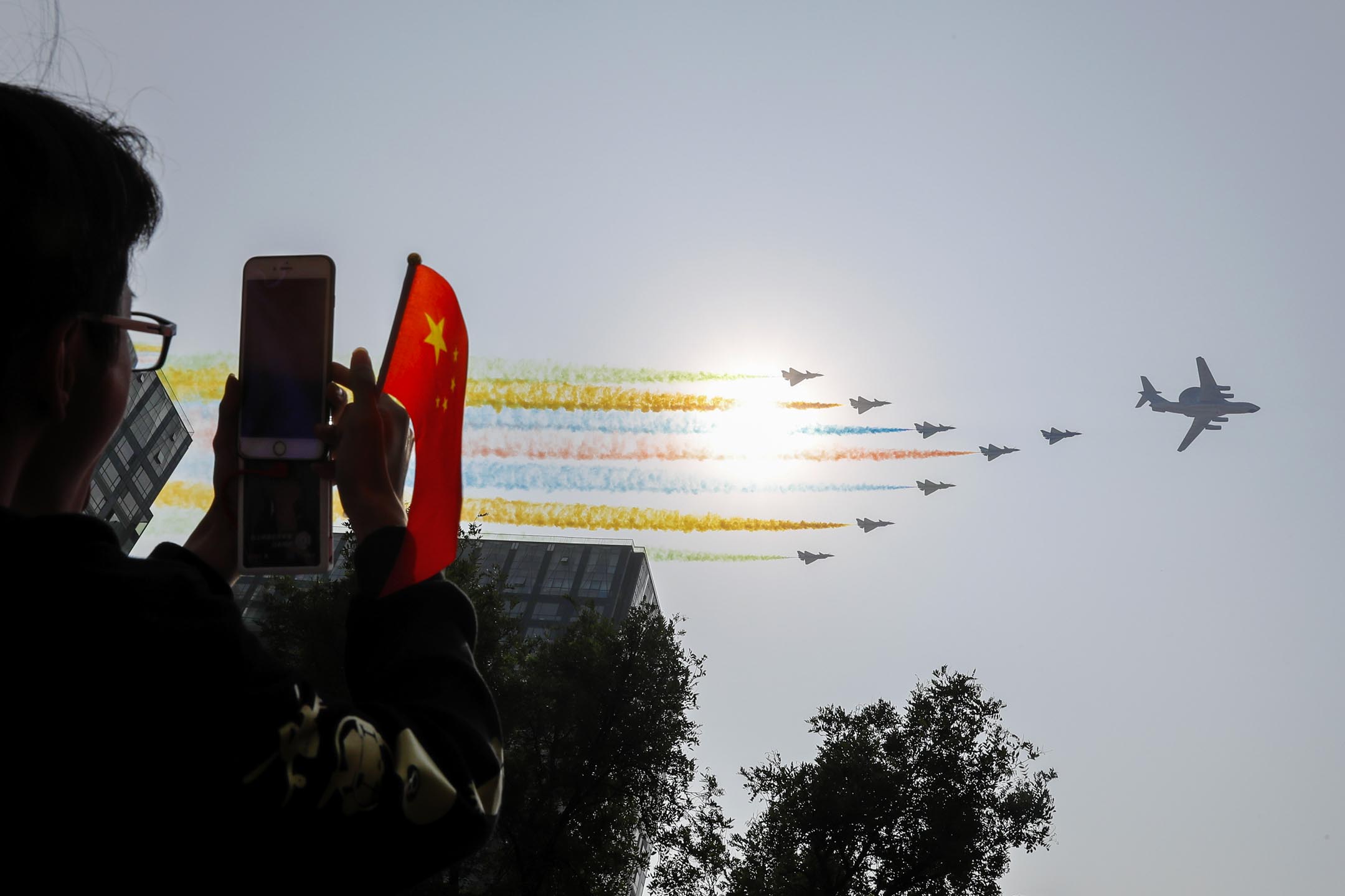 2019年10月1日,北京舉行的慶祝中國共產黨成立 70 週年的閱兵式上,一名男子用他的智能手機拍攝飛行中的中國軍機與彩色煙霧。