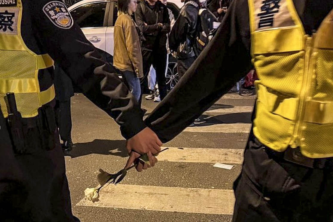 2022年11月27日凌晨,警察圍堵上海烏魯木齊中路聚集的大量市民,其中兩名警察共同手執一支白花。