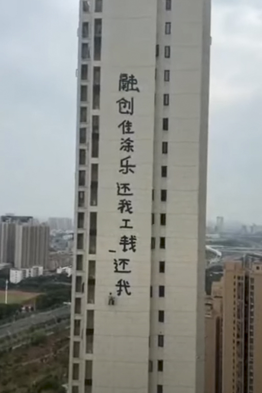 一位工人吊着繩索在一座住宅樓的外牆上刷標語,內容為「融創佳塗樂還我工錢,還我血汗錢」,結尾還畫了兩隻烏龜。